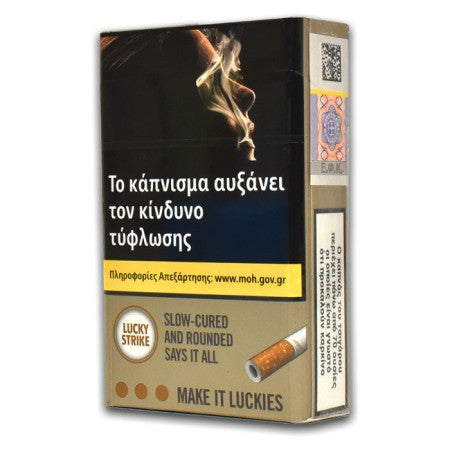 A pack of Lucky Strike cigarettes shown at a three-quarter angle on a plain white background. The front features a prominent dark panel with Greek warning text and a graphic of a smoking cigarette, while the lower section is beige with the Lucky Strike branding and a small illustration of a cigarette.